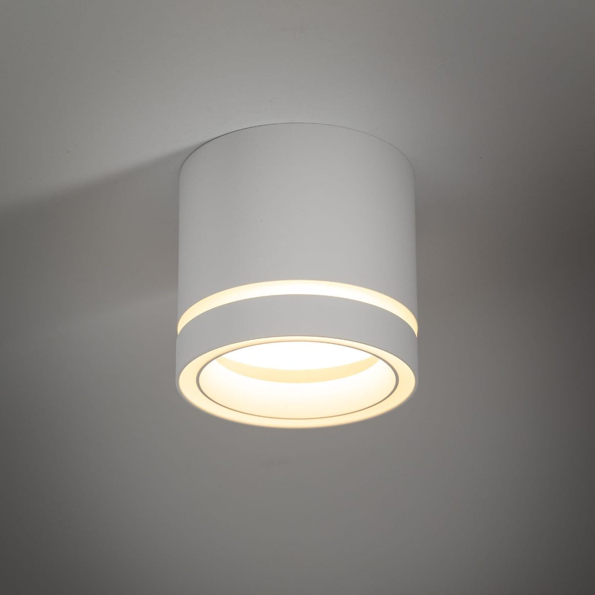 Lampa sufitowa Jet 1xGX53 w kolorze białym, okrągła, z delikatnymi żłobieniami wokół źródła światła. Ujęcie z dołu, ukazujące gładką powierzchnię i ciepłe światło emitowane z wnętrza. Montowana na suficie.