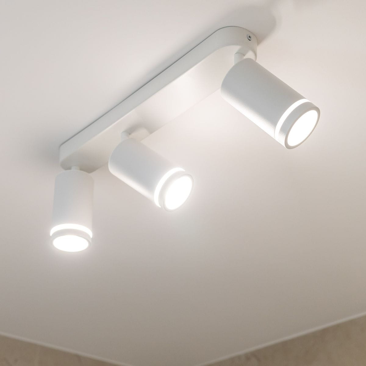 Lampa sufitowa Jet z trzema reflektorami GU10 w białej obudowie. Reflektory o cylindrycznym kształcie, zamontowane na prostokątnej podstawie. Ujęcie z dołu, ukazujące włączone źródła światła. Tło jasne, minimalistyczne.