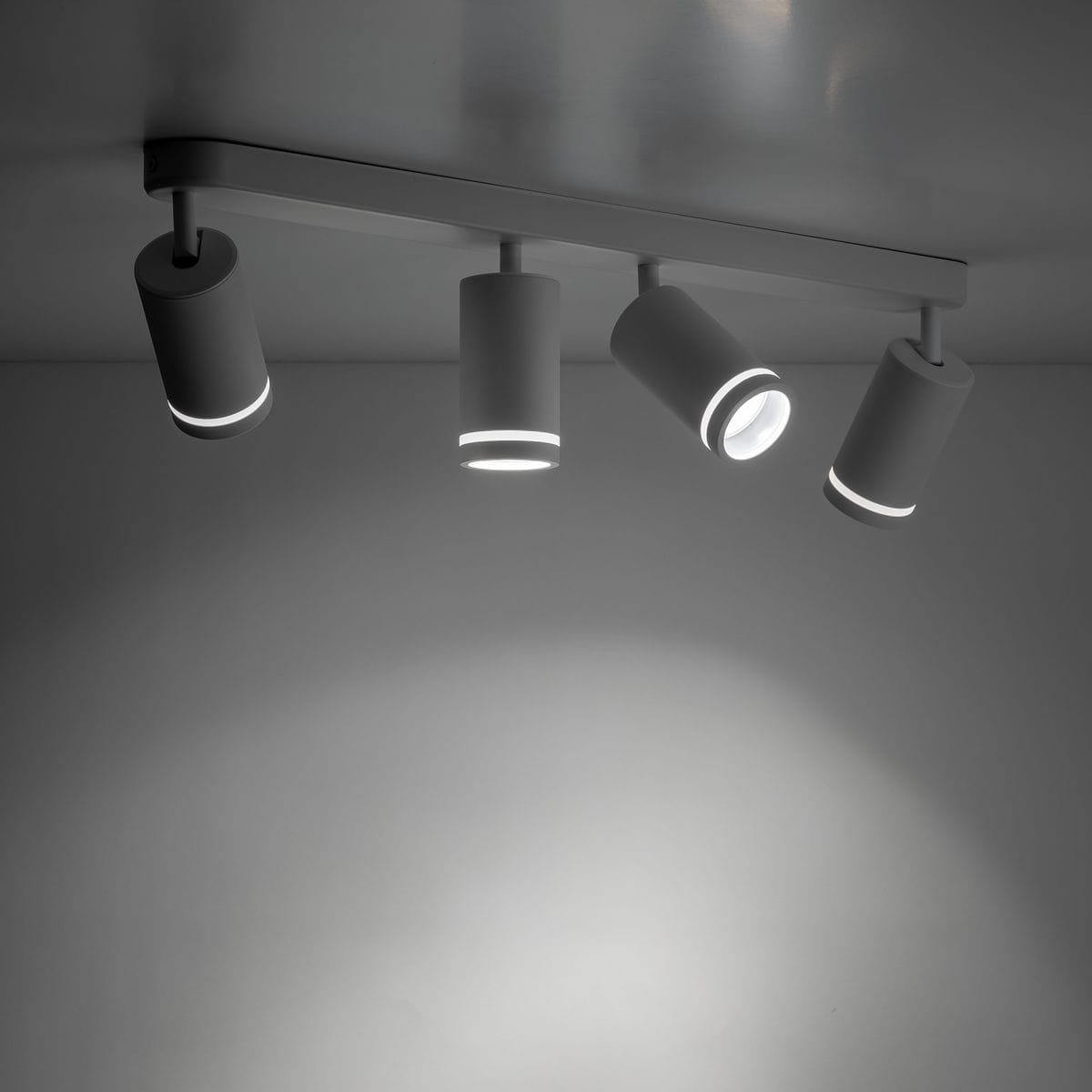 Lampa sufitowa Jet w kolorze białym, z czterema reflektorami GU10. Reflektory mają cylindryczny kształt i są zamontowane na prostokątnej listwie. Ujęcie z dołu, ukazujące rozproszone światło na tle szarym.