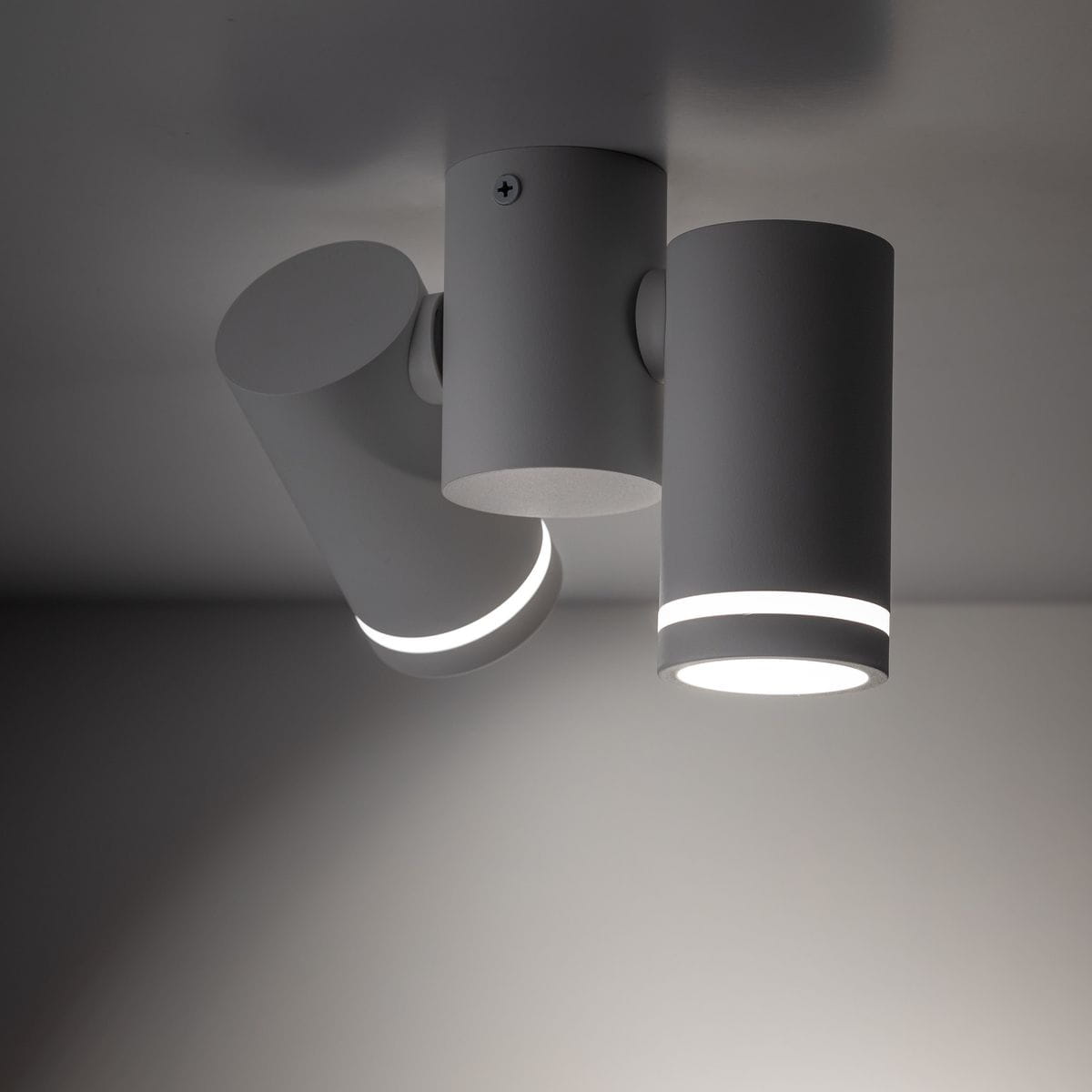 Lampa sufitowa Jet w białym kolorze, z dwoma spotami GU10. Ujęcie z dołu, pokazujące cylindryczne klosze w różnych pozycjach. Klosze mają wąskie pierścienie emitujące światło, montowane na okrągłej podstawie. Tło jednolite, szare.