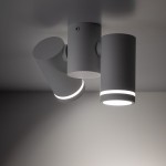 Lampa sufitowa Jet w białym kolorze, z dwoma spotami GU10. Ujęcie z dołu, pokazujące cylindryczne klosze w różnych pozycjach. Klosze mają wąskie pierścienie emitujące światło, montowane na okrągłej podstawie. Tło jednolite, szare.