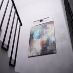 Kinkiet Gallery IP44 w kolorze czarnym z jedną żarówką LED 20W, zamontowany na ścianie, oświetlający abstrakcyjny obraz. Ujęcie z dołu, pokazujące kąt montażu oraz detale lampy. Tło w odcieniach szarości.