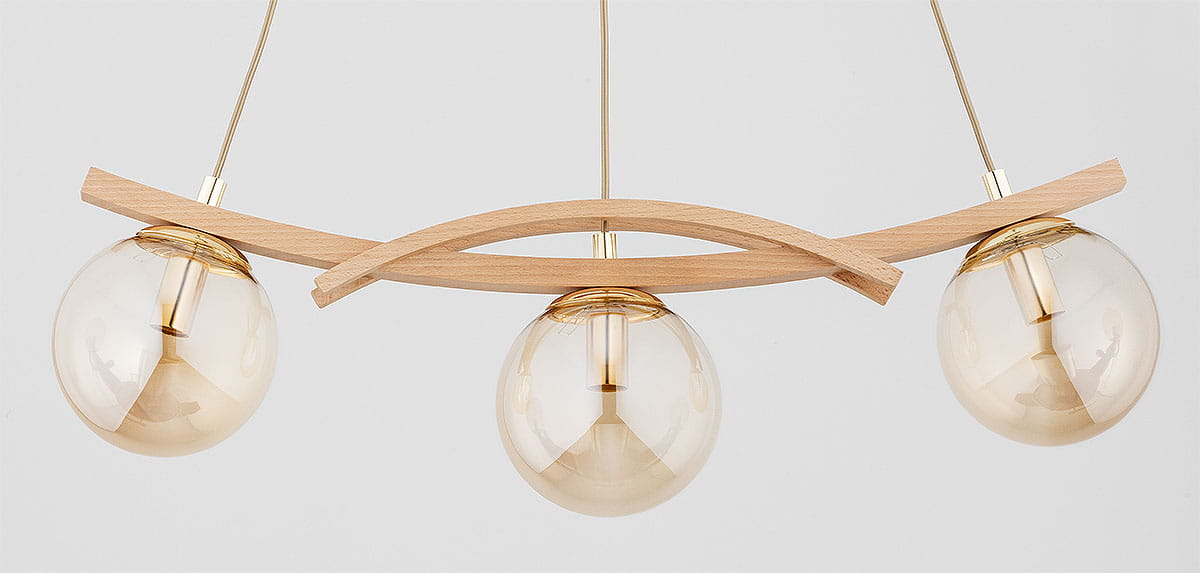 Lampa wisząca Sana z trzema kulistymi kloszami w kolorze złotym, zawieszona na drewnianym, łukowatym elemencie. Klosze wykonane z przezroczystego szkła, z widocznymi żarówkami E14. Całość w nowoczesnym stylu, na jasnym tle.