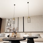 Czarna lampa wisząca Vander z metalową klatką i złotymi detalami, z jedną oprawą E27. Zawieszona na cienkim przewodzie, w nowoczesnym wnętrzu z kanapą i okrągłymi stolikami. W tle neutralne ściany i półka z dekoracjami.