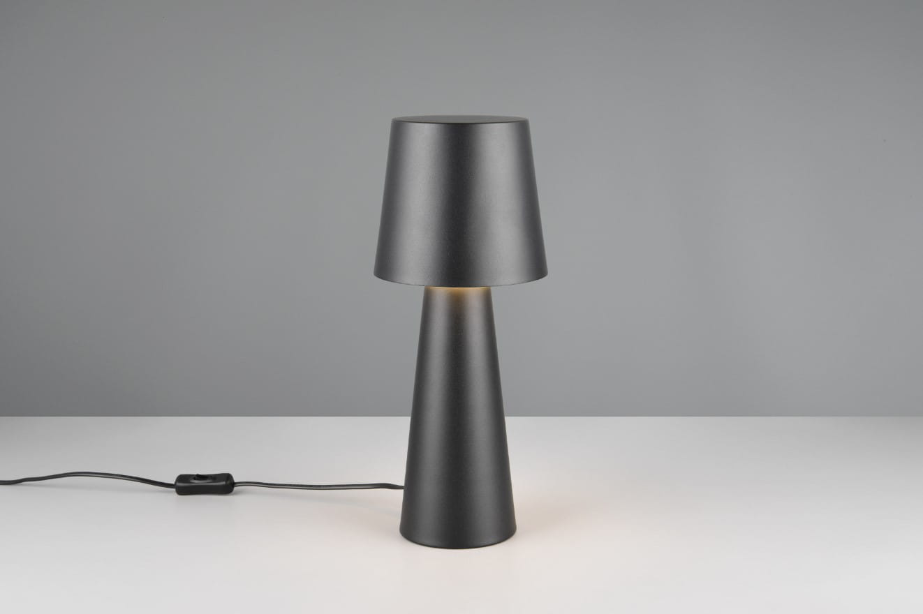 Czarna lampa stołowa Nubian z kloszem w kształcie odwróconego stożka, wykonana z matowego materiału. Stoi na białym blacie, z widocznym kablem i włącznikiem. Źródło światła E27. Tło w odcieniach szarości.