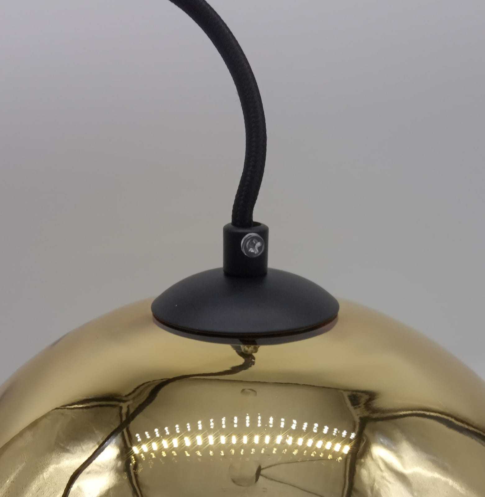 Lampa wisząca Mirror Glow D25 w kolorze złotym z metalowym kloszem. Widoczny czarny element montażowy oraz kabel tekstylny. Klosz o gładkiej, błyszczącej powierzchni, odbijający światło. Kąt ujęcia z góry, pokazujący detal mocowania.