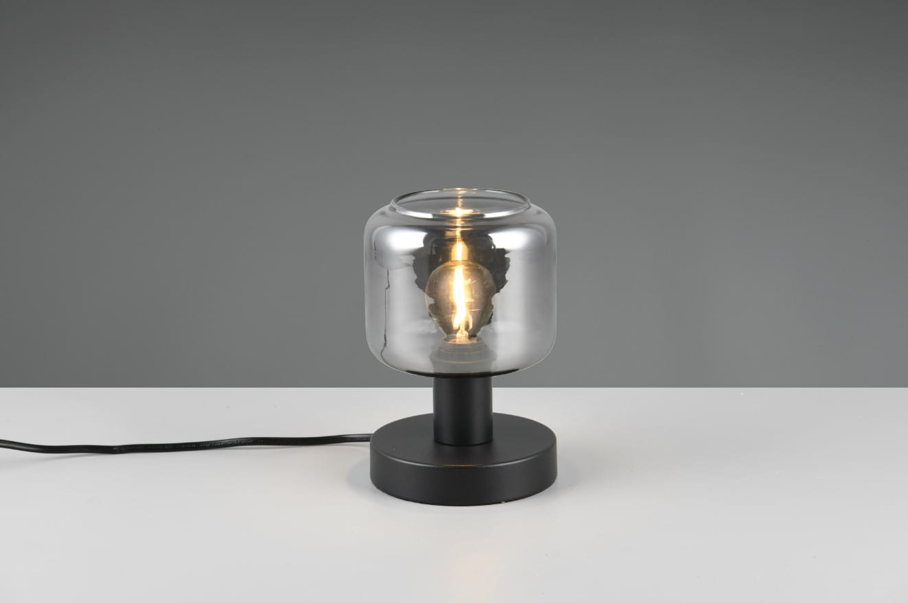 Lampa stołowa Mataro z czarną podstawą i przezroczystym kloszem, zamontowana na okrągłej podstawie. Widoczna żarówka E14, przyciągająca wzrok dzięki prostemu designowi. Kabel zasilający widoczny z boku, tło szare.