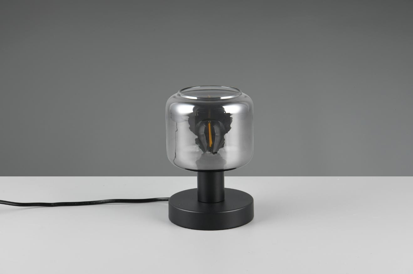 Czarna lampa stołowa Mataro z żarówką E14, z cylindrycznym kloszem z przezroczystego szkła. Ustawiona na okrągłej podstawie, z widocznym kablem zasilającym. Klosz odsłania źródło światła, tworząc nowoczesny akcent.