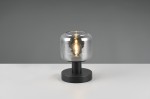 Lampa stołowa Mataro z oprawą w kolorze czarnym, z przezroczystym kloszem, stojąca na okrągłej podstawie. Źródło światła E14 widoczne wewnątrz klosza, o gładkiej fakturze. Tło w odcieniu szarym, produkt na białym blacie.