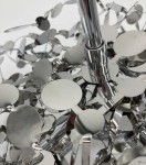 Lampa wisząca Monetti w kolorze chromu, z trzema oprawkami G9. Widoczne metalowe, błyszczące okręgi tworzące dekoracyjny wzór, z centralnym elementem montażowym. Zbliżenie na detale konstrukcji lampy.
