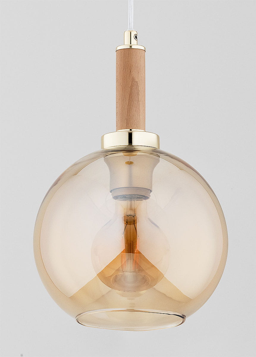 Lampa wisząca Hetty 1xE27 w złotym wykończeniu z drewnianym uchwytem. Okrągła, przezroczysta klosz w odcieniu bursztynowym, zawieszona na cienkim przewodzie. Widoczna żarówka wewnątrz klosza, montowana na suficie.