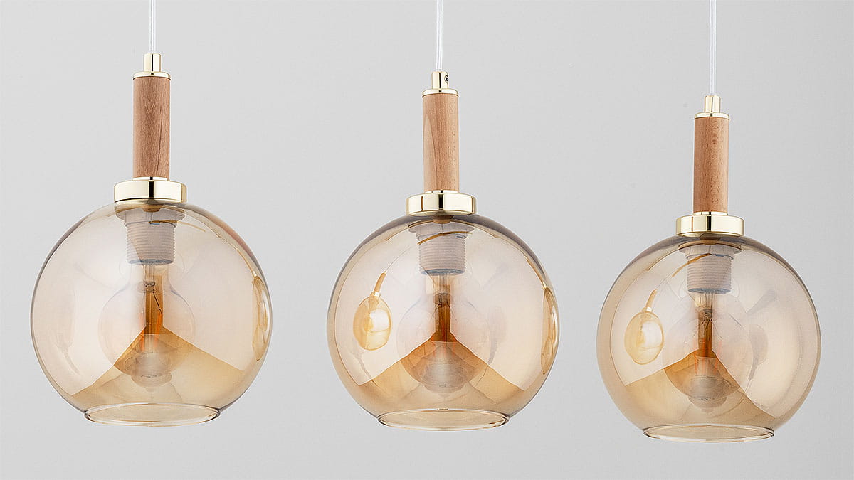 Lampa wisząca Hetty z trzema kloszami w kształcie kul, w kolorze złotym z drewnianymi elementami. Klosze wykonane z przezroczystego szkła w odcieniu brązowym, z widocznymi żarówkami E27. Ujęcie frontalne, lampy zawieszone na białych przewodach.