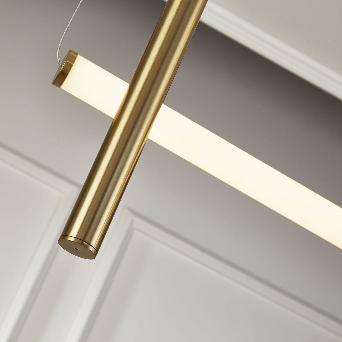 Lampa wisząca O-line L-4 w mosiężnym wykończeniu, z czterema źródłami LED, z prostokątnym kloszem w białym kolorze. Zbliżenie na metalowy element i klosz, zawieszony na cienkich linkach, na tle jasnej ściany.