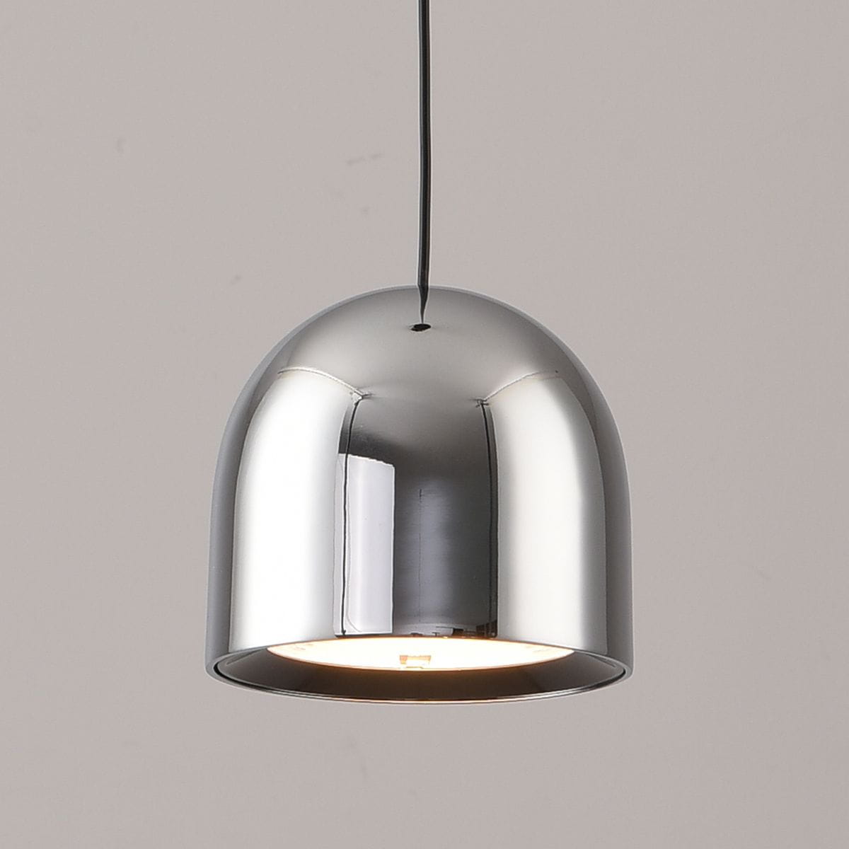 Lampa wisząca Petite w kolorze chromu z jedną żarówką LED o mocy 6W. Ma zaokrąglony kształt, zawieszona na cienkim przewodzie, z otwartą dolną częścią, przez którą wydobywa się światło. Tło w jasnym odcieniu.