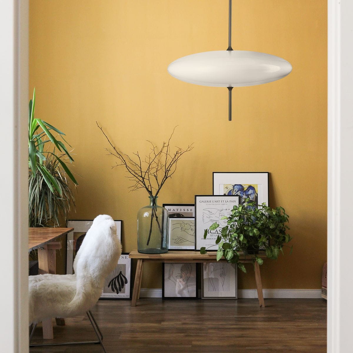 Lampa wisząca Piatto 3xE27 w kolorze czarnym i białym, o owalnym kształcie, zawieszona na cienkim kablu w jasnym wnętrzu z żółtymi ścianami, otoczona roślinami i dekoracyjnymi obrazami na ścianie.