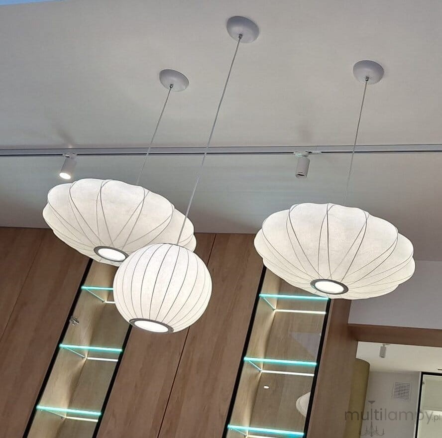 Lampa wisząca Silk D30 w kolorze białym, z jednym miejscem na żarówkę E27. Posiada trzy klosze w kształcie muszli, zwisające na cienkich linkach. Widoczna jest także sufitowa oprawa montażowa i tło z drewnianymi panelami.