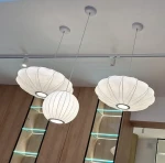 Lampa wisząca Silk D30 w kolorze białym, z jednym miejscem na żarówkę E27. Posiada trzy klosze w kształcie muszli, zwisające na cienkich linkach. Widoczna jest także sufitowa oprawa montażowa i tło z drewnianymi panelami.