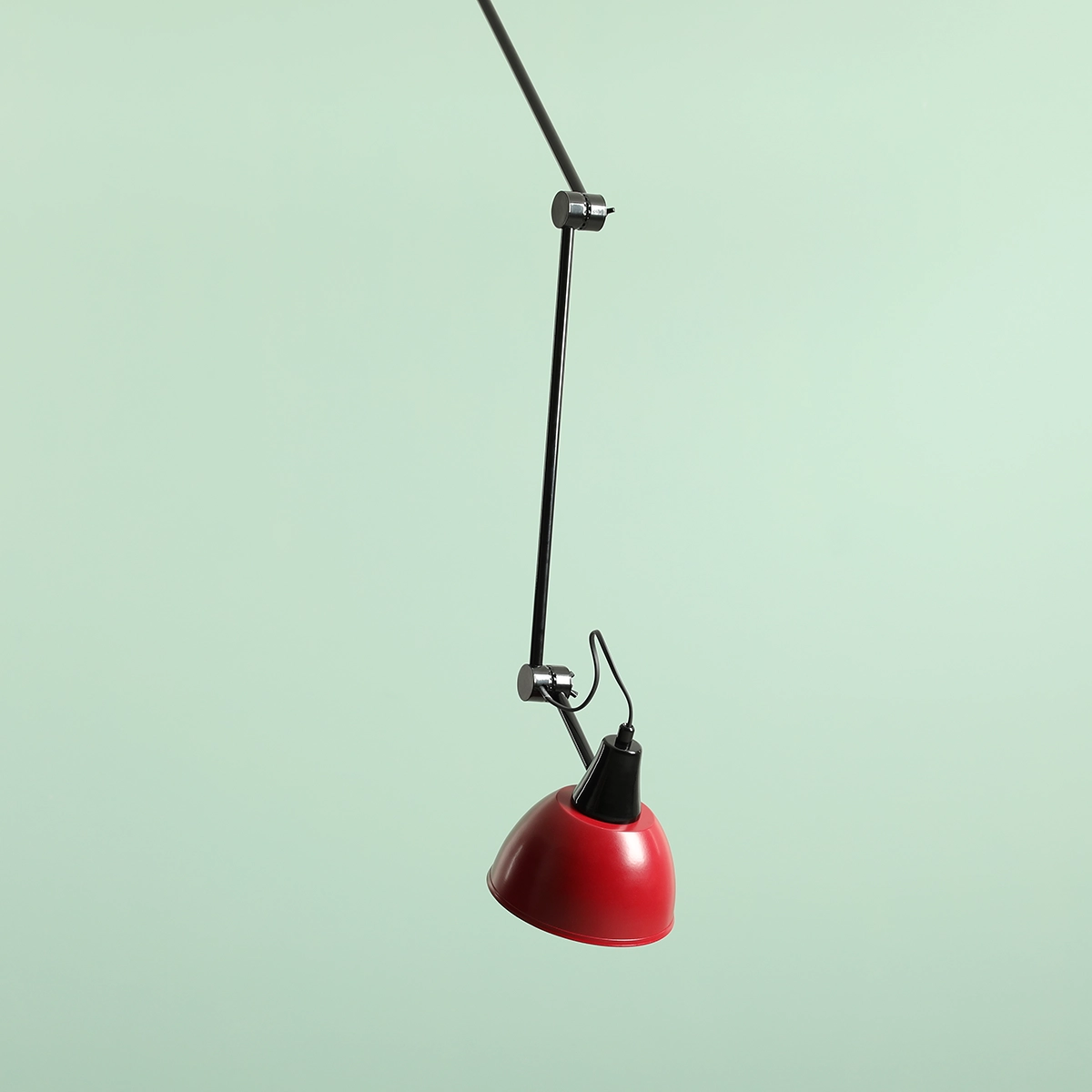 Czarna lampa wisząca Aida z czerwonym kloszem, zawieszona na cienkim, metalowym ramieniu. Klosz ma zaokrąglony kształt, a całość jest zamocowana do sufitu. Tło w jasnym odcieniu zieleni.