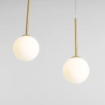 Lampa wisząca Bosso 4xE14 w kolorze mosiądzu, z dwiema okrągłymi kloszami z mlecznego szkła. Produkt zawieszony na cienkich linkach, z minimalistycznym designem, na jednolitym tle.