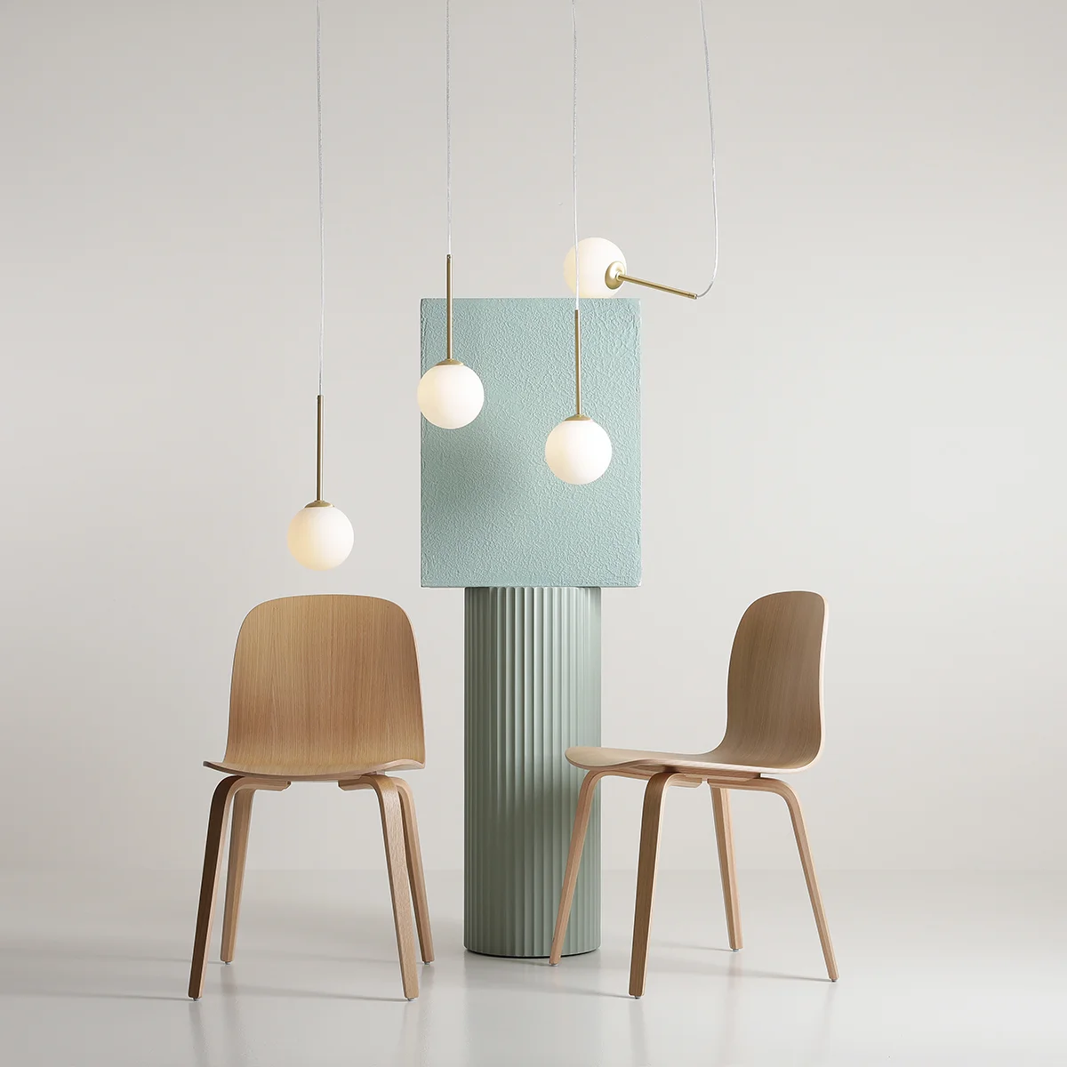 Lampa wisząca Bosso 4xE14 w mosiężnej oprawie, z czterema białymi, okrągłymi kloszami, zawieszona nad zielonym, teksturowanym słupem. W tle dwa drewniane krzesła, na jasnym tle, w minimalistycznej aranżacji.