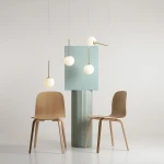 Lampa wisząca Bosso 4xE14 w mosiężnej oprawie, z czterema białymi, okrągłymi kloszami, zawieszona nad zielonym, teksturowanym słupem. W tle dwa drewniane krzesła, na jasnym tle, w minimalistycznej aranżacji.