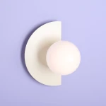 Kinkiet Loop Cream z białą, półokrągłą osłoną i okrągłą, mleczną żarówką. Zamontowany na jasnofioletowej ścianie, prezentuje nowoczesny design. Widoczny detal to gładka faktura materiału.