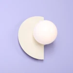 Kinkiet Loop Cream 1xE14 w kształcie półokręgu z białą, matową kulistą żarówką. Ściana w tle w odcieniu lawendowym. Kinkiet zamontowany na ścianie, z minimalistycznym designem, podkreślającym nowoczesny styl.