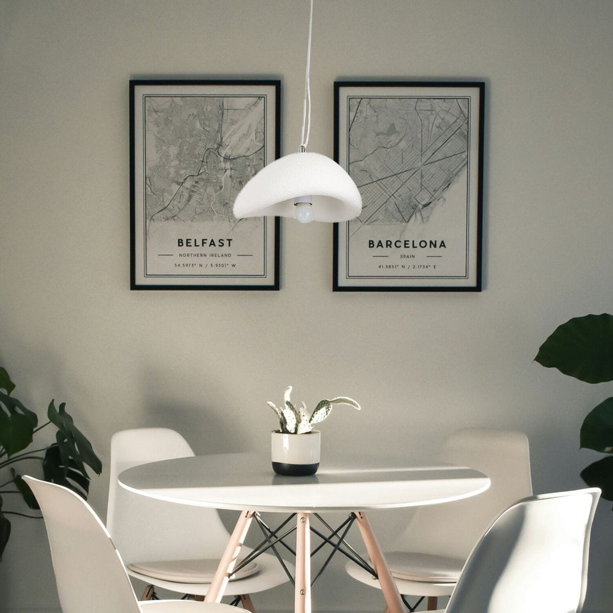 Lampa wisząca Stone D30 w kolorze białym, z jedną oprawą E27, o zaokrąglonym kształcie. Zawieszona nad okrągłym stołem, w nowoczesnym wnętrzu, w towarzystwie rośliny doniczkowej i plakatów na ścianie.
