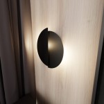 Kinkiet Form 9 w kolorze czarnym, o okrągłym kształcie z półotwartą strukturą, montowany na jasnej drewnianej ścianie. Emituje miękkie światło, zbliżenie na detal oświetlenia oraz teksturę powierzchni.