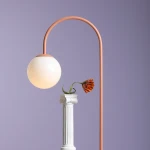 Lampa stołowa Una Coral z abażurem w kształcie kuli w odcieniu mlecznym, na różowym, giętym stelażu. Ustawiona obok białego kolumny z kwiatem. Tło w delikatnym fioletowym odcieniu.