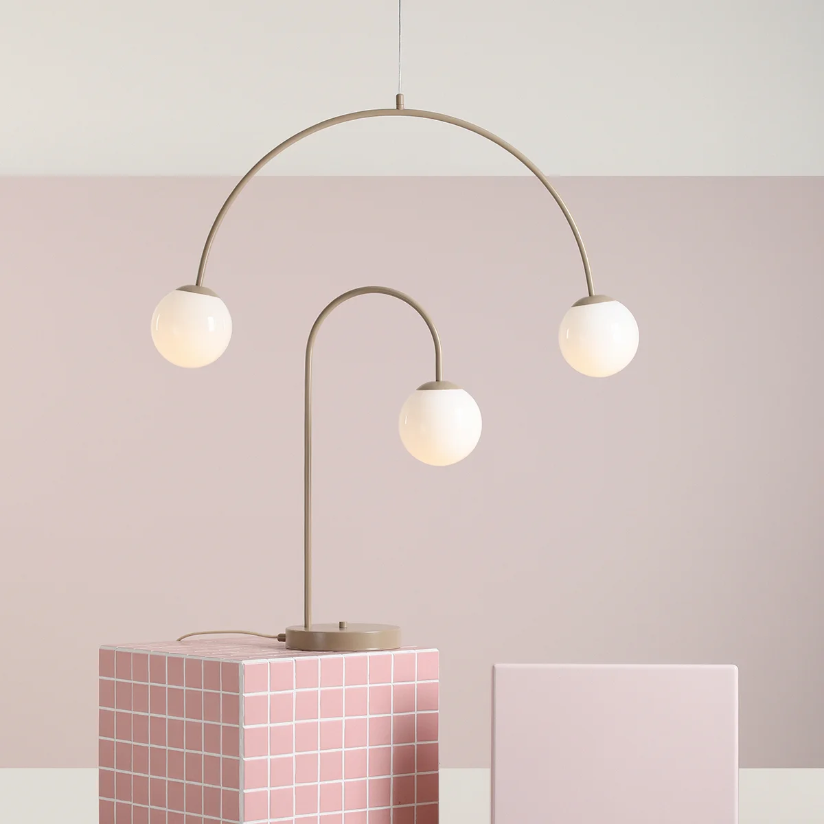 Lampa wisząca Una Beige z dwiema kulistymi, białymi kloszami na łukowatych ramionach w kolorze beżowym. Produkt zawieszony nad różowym tłem, na podwyższeniu w formie kwadratowego słupa z kafelkami.