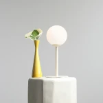 Kremowa lampa stołowa Pinne z okrągłym, matowym kloszem na cienkiej nodze, umieszczona obok żółtej wazonu z zieloną rośliną. Produkt stoi na stołku o okrągłej podstawie, w neutralnym, szarym tle.