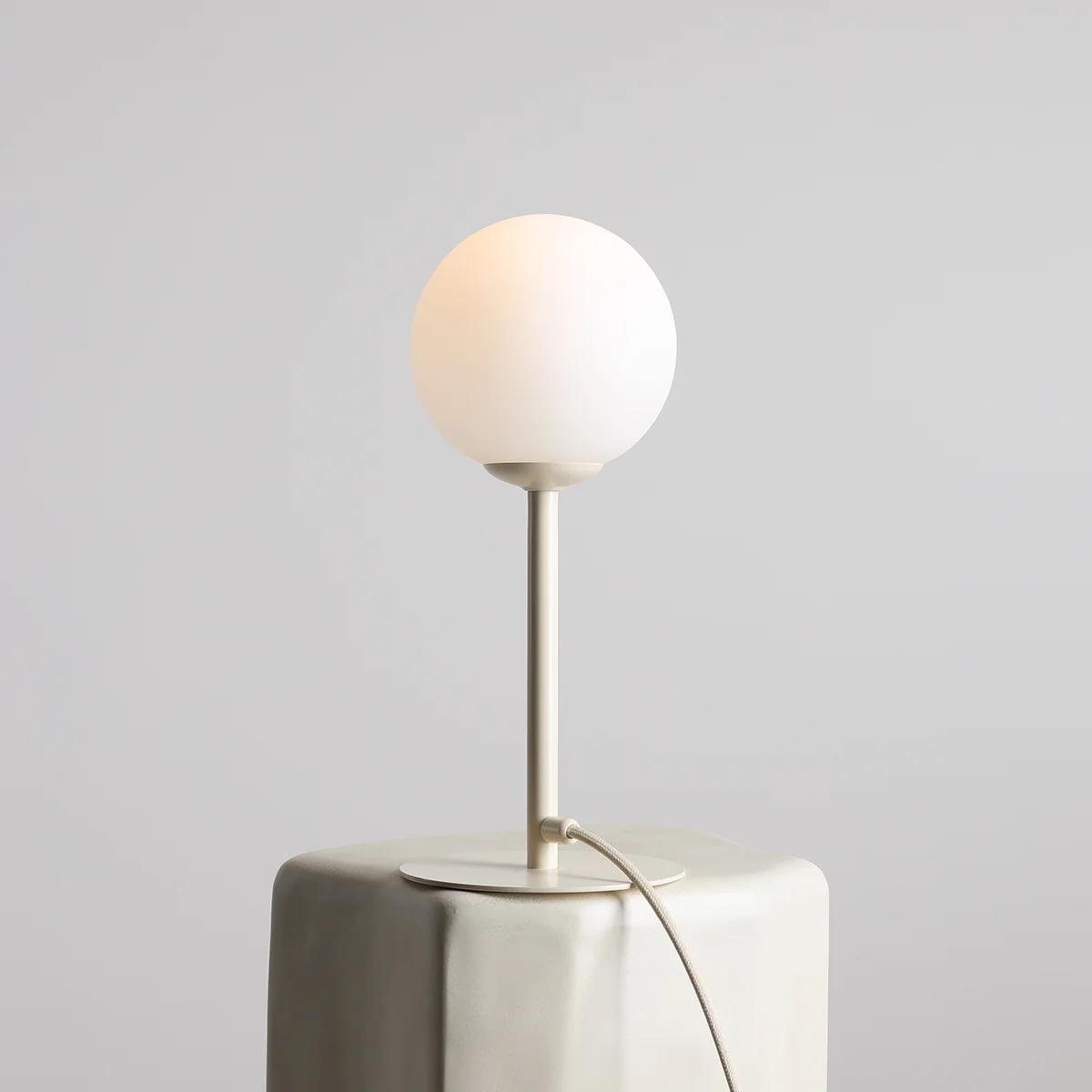 Kremowa lampa stołowa Pinne z kulistym kloszem z matowego szkła, osadzona na cienkim, metalowym stelażu. Lampa stoi na prostokątnej podstawie, z widocznym przewodem. Tło w odcieniu szaro-beżowym.