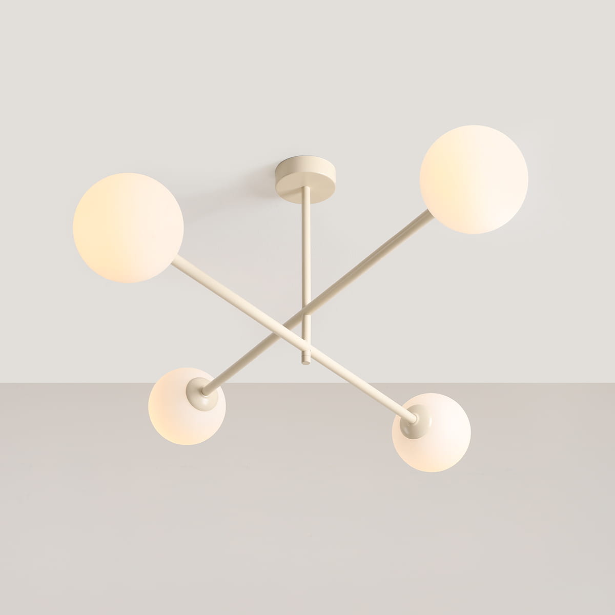 Kremowa lampa sufitowa Pinne z czterema oprawami E14, o nowoczesnym designie, z okrągłymi kloszami w kolorze mlecznym, umieszczona na krzyżujących się metalowych ramionach. Ujęcie z dołu, na jasnym tle.