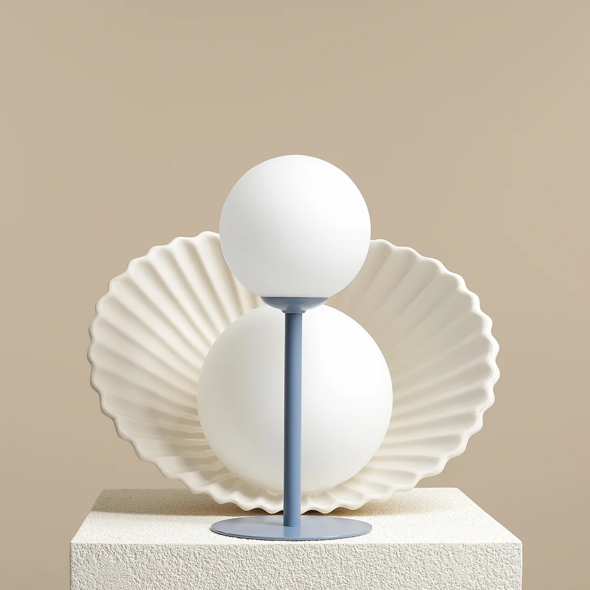 Lampa stołowa Pinne 1xE14 w kolorze niebieskim, z okrągłym białym kloszem na smukłej podstawie, umieszczona przed dekoracyjnym tłem w kształcie muszli. Produkt stoi na kwadratowej, teksturowanej powierzchni.