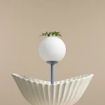 Lampa stołowa Pinne z niebieskim trzonkiem, z białą, okrągłą kloszem i ozdobnym zielonym elementem roślinnym na górze. Ustawiona na ceramicznej podstawie w kształcie muszli, na jednolitym, jasnym tle.