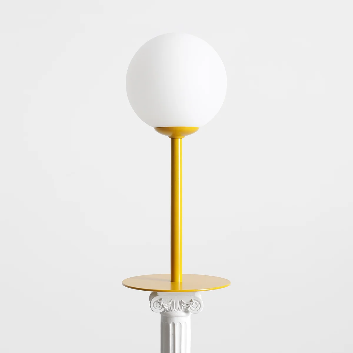 Lampa stołowa Pinne z żółtą podstawą i białym, okrągłym kloszem. Ustawiona w pozycji stojącej na okrągłej podstawie. Prosta forma, metalowy stelaż, elegancka i nowoczesna estetyka. Tło w kolorze białym.