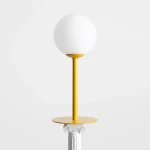 Lampa stołowa Pinne z żółtą podstawą i białym, okrągłym kloszem. Ustawiona w pozycji stojącej na okrągłej podstawie. Prosta forma, metalowy stelaż, elegancka i nowoczesna estetyka. Tło w kolorze białym.