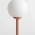 Lampa stołowa Pinne z koralowym stelażem i białym, okrągłym kloszem. Ujęcie z boku, pokazujące detale podstawy i klosza. Prosta, nowoczesna forma, idealna do aranżacji wnętrz.