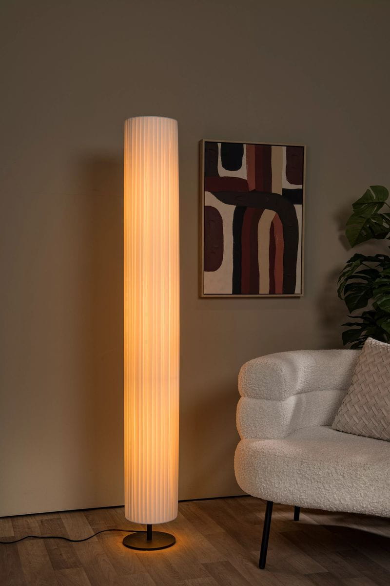 Lampa podłogowa Fold 3xE27 w kolorze czarno-białym, o cylindrycznym kształcie, z białą, teksturowaną obudową. Ustawiona w rogu pokoju obok jasnej sofy, emitująca ciepłe światło. Widoczny czarny stelaż i kabel zasilający. Tło w neutralnym odcieniu.
