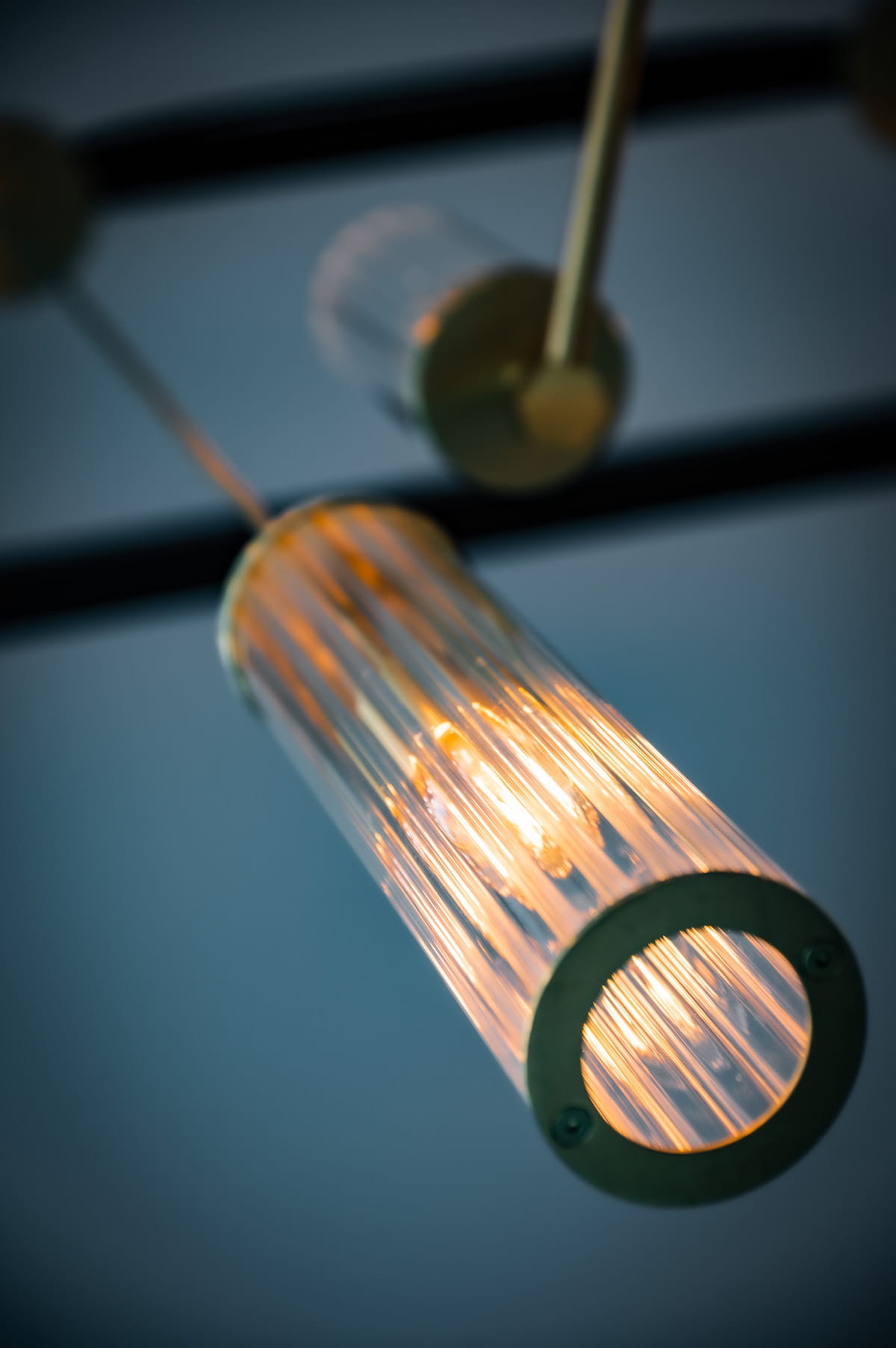 Lampa wisząca Hampton z metalowym elementem w kolorze mosiądzu. Zawiera szklany klosz w formie cylindrycznej, z wyraźnymi liniami, emitujący ciepłe światło. Ujęcie z dołu, pokazujące detal klosza i źródło światła.