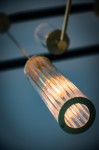 Lampa wisząca Hampton z metalowym elementem w kolorze mosiądzu. Zawiera szklany klosz w formie cylindrycznej, z wyraźnymi liniami, emitujący ciepłe światło. Ujęcie z dołu, pokazujące detal klosza i źródło światła.