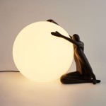 Lampa stołowa Woman-1 z czarną figurą siedzącą, obejmującą dużą, okrągłą, białą kulę. Materiał matowy, a kształt lampy nadaje jej nowoczesny charakter. Ujęcie z boku, na jasnym tle. Wymagana żarówka E27.