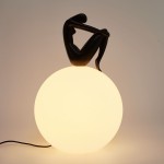 Lampa stołowa Woman-2 z czarną figurką siedzącą na mlecznobiałej, okrągłej podstawie. Figura przedstawia sylwetkę kobiety w pozycji siedzącej. Produkt z kablem zasilającym widocznym z boku.