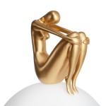 Lampa stołowa Woman-2 z podstawą w kształcie białej kuli, na której siedzi figura kobiety w złotym kolorze. Figura ma smukłą sylwetkę, z rękami owiniętymi wokół kolan, w pozycji siedzącej. Kąt ujęcia: z boku.