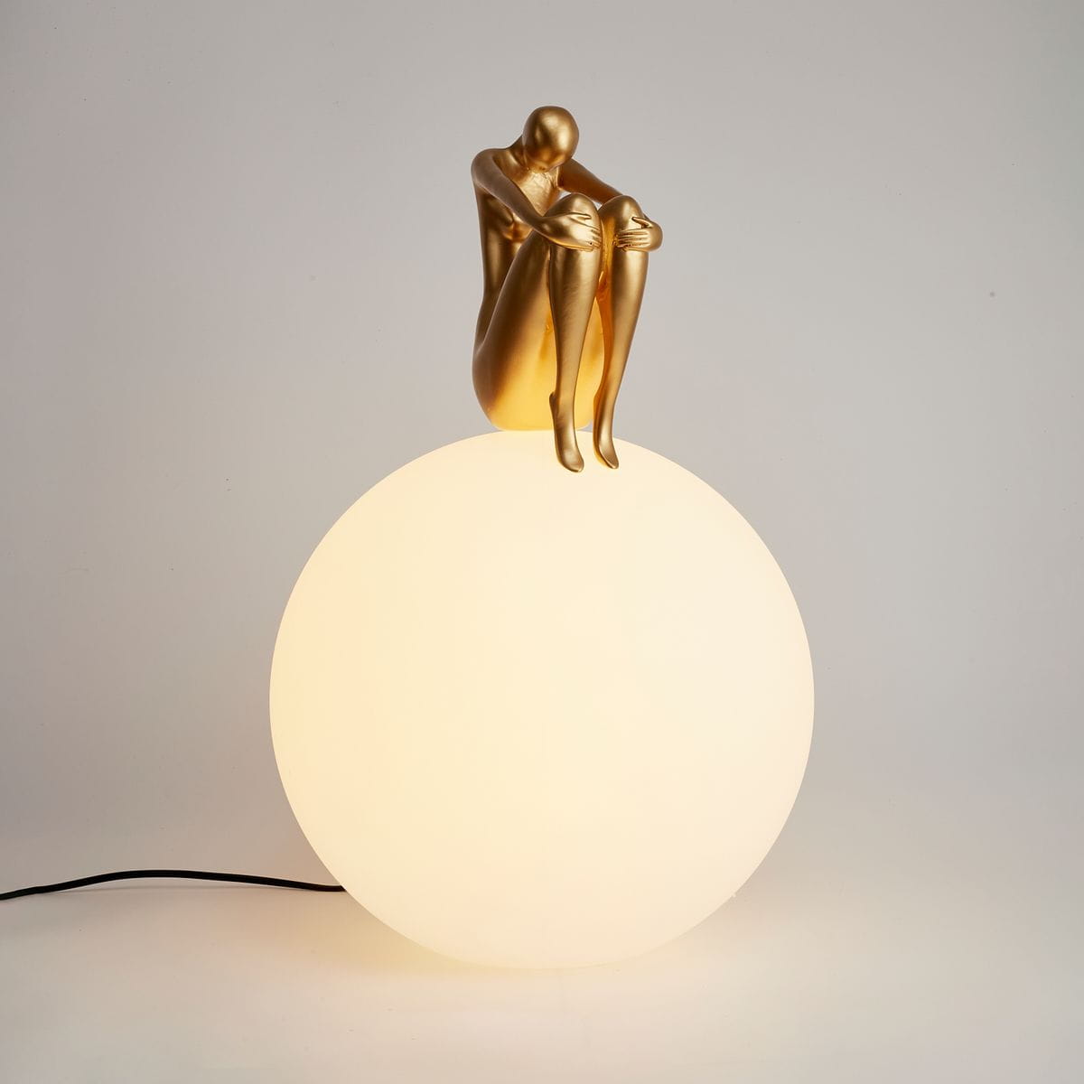 Lampa stołowa Woman-2 z złotą figurką siedzącą na matowej, białej kuli. Figurka ma smukłą sylwetkę, a lampa jest podłączona do zasilania przez czarny kabel. Tło jest jasne, co podkreśla elegancki design.