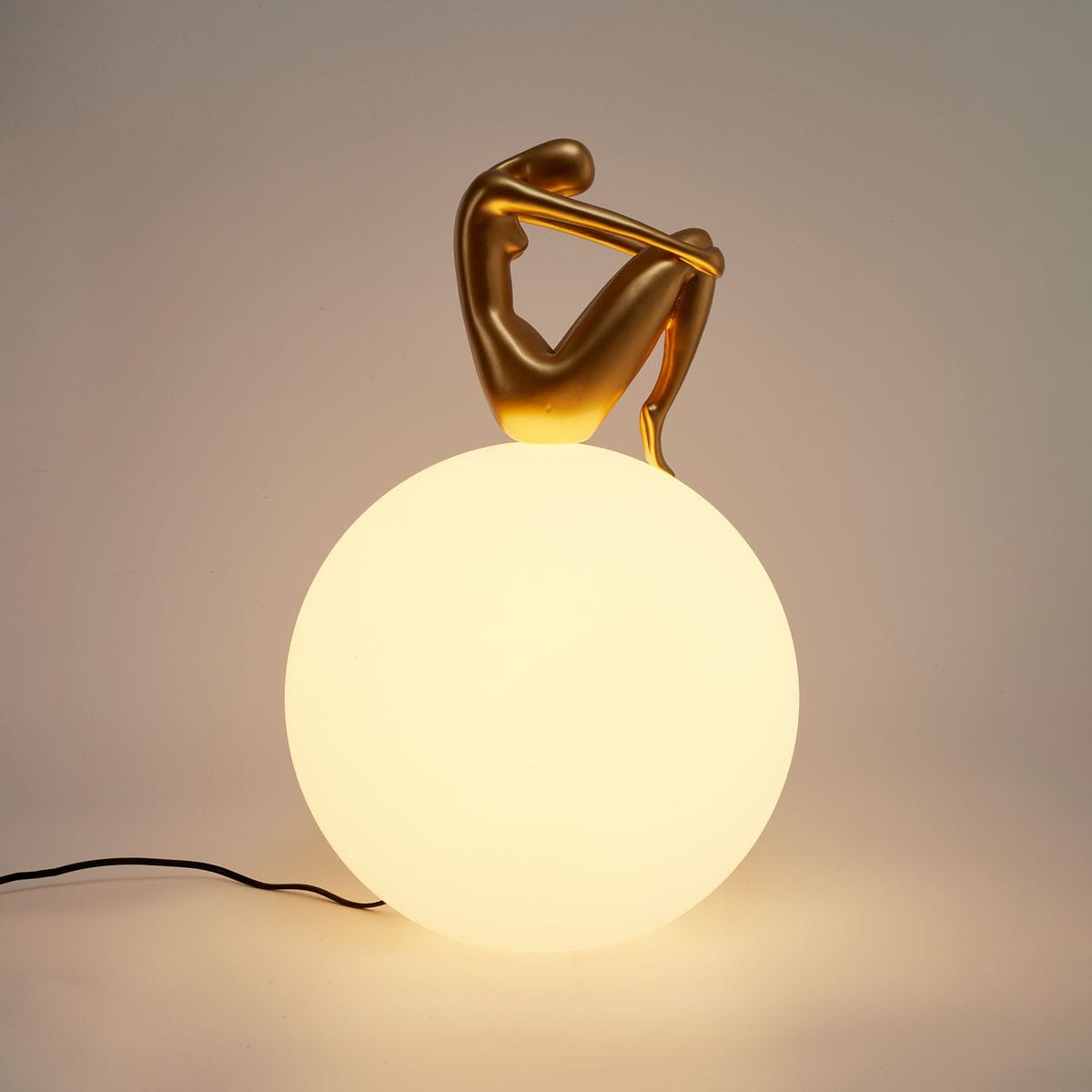 Lampa stołowa Woman-2 z złotą figurką siedzącą na dużej, matowej, białej kuli. Kąt ujęcia frontalny, widoczny kabel zasilający. Figurka ma zaokrąglone kształty, podkreślające elegancki design lampy.