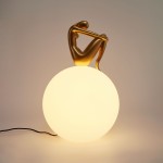 Lampa stołowa Woman-2 z złotą figurką siedzącą na dużej, matowej, białej kuli. Kąt ujęcia frontalny, widoczny kabel zasilający. Figurka ma zaokrąglone kształty, podkreślające elegancki design lampy.