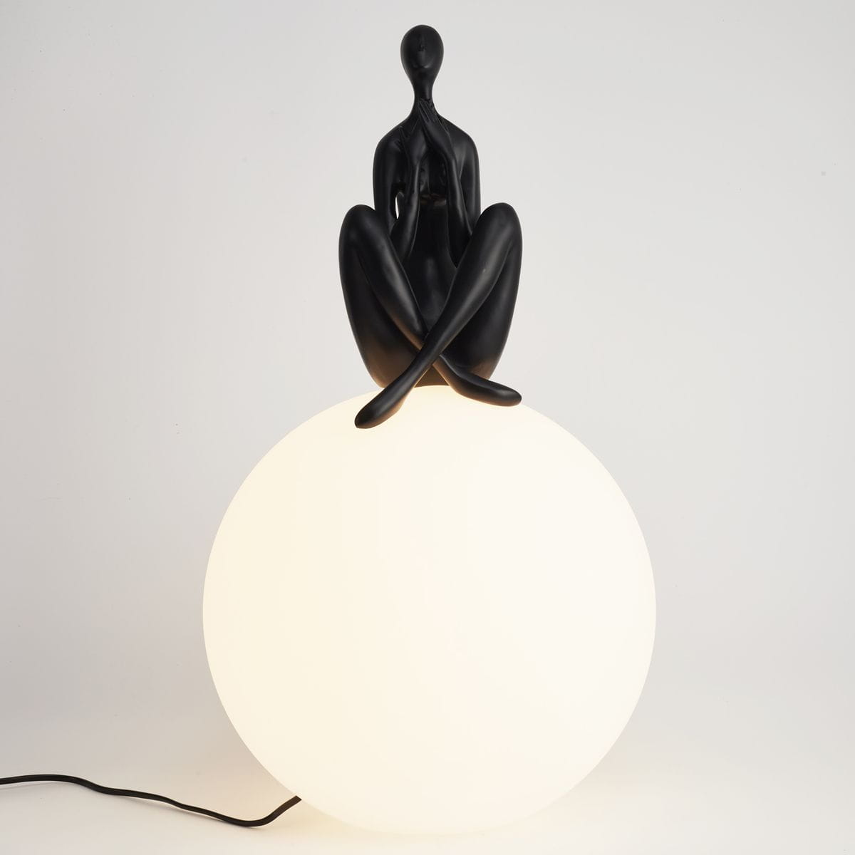 Lampa stołowa Woman-3 z czarną figurką siedzącą na białej, okrągłej podstawie. Figurka ma gładką, matową powierzchnię, a lampa jest podłączona do zasilania przez czarny kabel. Produkt prezentowany na jasnym tle.