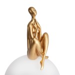 Lampa stołowa Woman-3 z figurką w złotym kolorze, siedzącą na białej, półprzezroczystej podstawie. Figurka ma gładką powierzchnię, z wyraźnymi detalami ciała, przyciągającą wzrok elegancją. Złącze E27 dla jednego źródła światła.